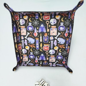 Könnte beinhalten: Ein schwarzes, quadratisches Würfeltablett mit einem sich wiederholenden Muster aus Zaubertränken, Totenköpfen, Schlüsseln, Motten und Knochen. Das Tablett hat erhöhte Kanten und vier gebogene Ecken. Das Design ist in Lila-, Gelb- und Weißtönen gehalten.