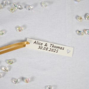 Può includere: Un'etichetta color crema con i nomi "Alice & Thomas" e la data "30.08.2025" stampati sopra, insieme a un piccolo simbolo a forma di cuore. Un nastro dorato è attaccato all'etichetta. Cristalli iridescenti sono sparsi intorno.