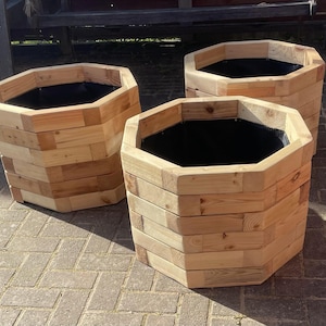 May include: Three hexagonal wooden planters. Each planter is made from stacked, light-coloured wooden planks. The interior of each planter is lined with a black material. The planters are arranged on a brick surface.