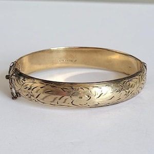 Può includere: Un braccialetto rigido color oro con un motivo floreale inciso. Il braccialetto a cerniera ha una catena di sicurezza e una chiusura. La superficie è incisa con un disegno dettagliato di fiori e foglie.