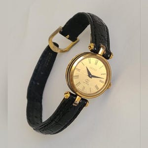 May include: A gold-toned Gucci watch with a round face and black leather band. The watch face features gold hands and Roman numeral hour markers. The band is a textured black leather with a gold buckle.