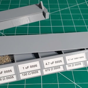 May include: A gray plastic parts organizer with multiple compartments, each labeled with component values like "1 uF 0805" and "4.7K Ω 0805." One compartment contains small, metallic components. The lid is partially open, revealing the contents.