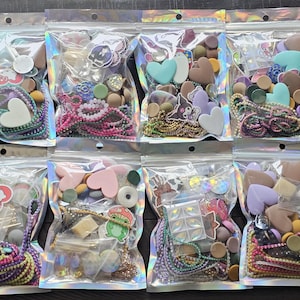 May include: Eight clear holographic resealable bags filled with assorted craft supplies including colorful beads, heart-shaped charms, small resin figures, metallic ball chains, and various flat-backed cabochons in a wide range of pastel and vibrant colors.