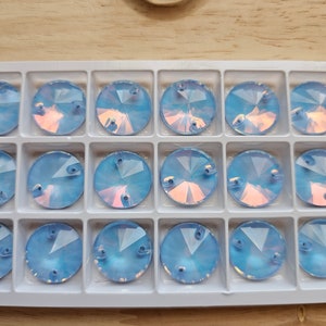 May include: A close-up of a white plastic container filled with 16 iridescent blue round rhinestones. The rhinestones are arranged in a grid pattern.