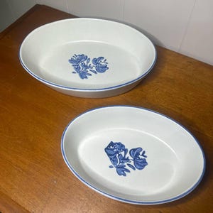 Puede incluir: Dos fuentes ovaladas. Las fuentes son blancas con un diseño floral azul en el centro y un borde azul. Las fuentes son de cerámica y están sobre una superficie de madera.