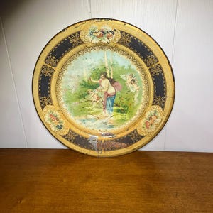 May include: Round vintage metal tray with a painted scene of a woman and cherubs in a garden. The tray has a black border with gold filigree and floral accents. The center image is framed with a gold border.