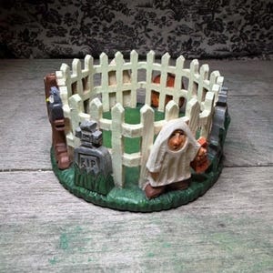 Op de afbeelding: Een cirkelvormig decoratief object met een wit lattenhek dat een groene basis omsluit. De scène bevat een grijze grafsteen met de letters RIP, een figuur in een wit spookkostuum en een bruin boomstamelement.