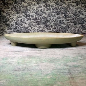 May include: A pale green, oval ceramic serving dish, supported by three small feet. The dish has a raised edge and a matte finish. The background displays a black and white floral pattern.