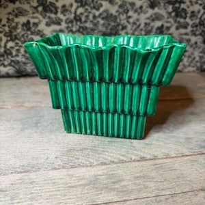 Op de afbeelding: Een levendige smaragdgroene keramische plantenbak met een getrapt ontwerp en verticale ribbels. De vierkante pot heeft een brede, uitlopende bovenrand die naar beneden toe smaller wordt, afgewerkt met een glanzende, reflecterende glazuurlaag.