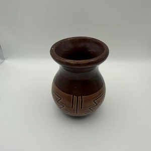 May include: A brown ceramic vase with a wide, rounded body and a narrow neck. The vase features a darker brown rim and a lighter brown band with a geometric design around the middle. The vase is approximately 15 cm tall.