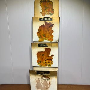 Könnte beinhalten: Vintage Wandorganizer mit vier Fächern. Der obere Bereich und jedes Fach weisen dekorative Illustrationen von Küchenutensilien und Obst auf. Die Fächer sind mit "LETTERS", "MEMO" und "MISC" beschriftet. Der Organizer hat eine verwitterte, goldfarbene Oberfläche.