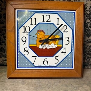 May include: A square wall clock with a light brown wooden frame. The clock face features a blue background with white dots, an octagonal center, black numbers, and a graphic of a yellow hen sitting on white eggs. The text Elgin Quartz is visible.
