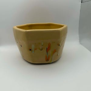 May include: A ceramic, hexagonal-shaped bowl in a light tan color. The bowl has a smooth exterior with colorful drips of orange, yellow, and black paint. The interior of the bowl is a darker yellow.