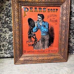 Op de afbeelding: Een vintage-stijl reclame voor Pears' Soap ingelijst in gesneden hout. De oranje print toont een persoon die zich scheert voor een spiegel met de tekst: Pears' Soap, A Luxury for Shaving, Pears Shaving Soap, 12 Months of Comfort for 12 Pence.