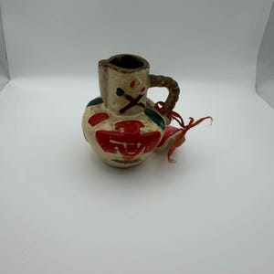 May include: A small, hand-painted ceramic jug with a rounded body and a handle. The jug is off-white with red, green, and black painted details. A red ribbon is tied to the handle. The jug has a rustic, folk art aesthetic.