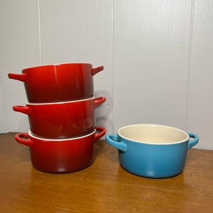 May include: A stack of three red ceramic soup bowls with handles, next to a single blue bowl. The bowls have a glossy finish and a white interior. The bowls are on a wooden surface.