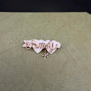 Puede incluir: Broche blanco y dorado con el nombre "Amy" escrito con alambre dorado. El broche tiene un diseño en forma de corazón con un dije de estrella colgante. El fondo es de tela verde claro.