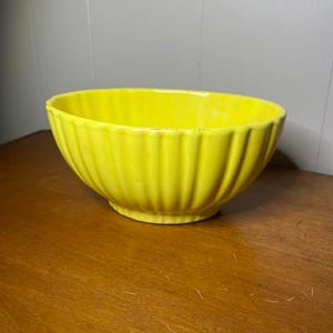 Puede incluir: Un tazón de cerámica amarillo brillante con un diseño acanalado. El tazón está vacío y se encuentra sobre una superficie de madera.