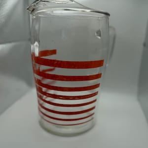 May include: A clear glass pitcher with a handle and a spout. The pitcher is decorated with horizontal red stripes. The pitcher is empty and appears to be vintage.