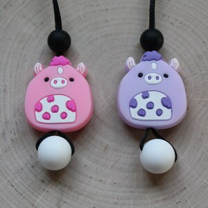 Può includere: Due morsi di silicone a forma di mucca con macchie bianche e nere. Uno è rosa e l'altro è viola. Ogni morso ha una perla di silicone bianca attaccata alla parte inferiore.