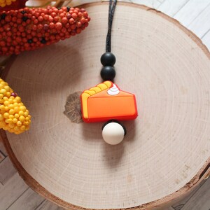 May include: A silicone necklace with a pumpkin pie slice charm, black beads, and a white bead. The necklace is on a wooden surface.