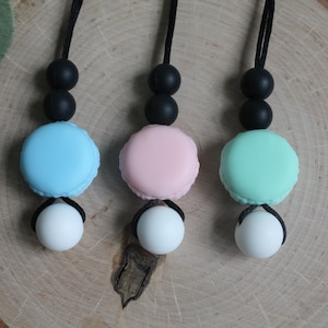 Pode incluir: Três colares de silicone com pingentes de macarons. Os macarons são azuis, rosa e verdes. Cada colar tem duas contas de silicone pretas e uma conta de silicone branca.