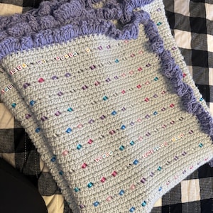 May include: A light blue crocheted blanket with a purple crocheted border. The blanket is decorated with colorful beads.