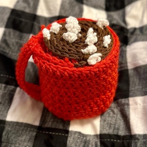 May include: A red crocheted mug filled with brown yarn and white yarn resembling hot chocolate with marshmallows.