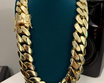 May include: A gold-colored chain necklace with large, interlocking links. The necklace is displayed on a teal mannequin. The clasp is visible, and the links reflect light, creating a shiny appearance. The necklace is a statement piece.