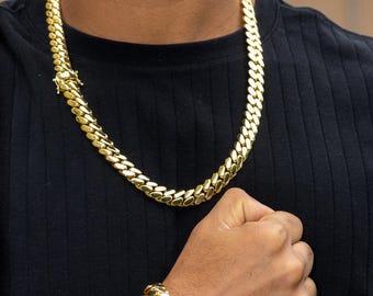 May include: A gold-colored chain necklace and matching bracelet. The jewelry features a thick, curb link design. The necklace is around the neck, and the bracelet is on the wrist. The person is wearing a black shirt.