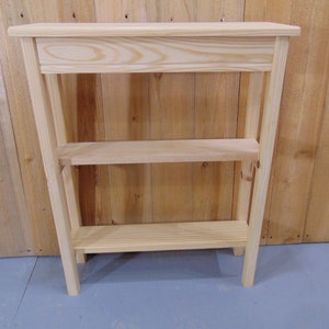 May include: A natural wood console table with two shelves. The table is made of unfinished pine wood and has a simple, rustic design.