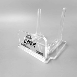 Könnte beinhalten: Ein transparenter Acryl-Ständer für eine Vintage-Handheld-Spielkonsole Atari Lynx. Der Ständer trägt den Text "TYPE ATARI LYNX" und "RELEASE 1989".
