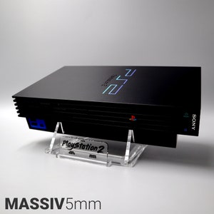 Peut inclure: Une console PlayStation 2 noire avec un support en acrylique transparent. Le support porte l'inscription "PlayStation 2". La console est posée sur une surface blanche.