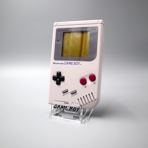 Könnte beinhalten: Eine weiße Nintendo Game Boy Handheld-Spielkonsole mit einem grauen Bildschirm und roten Knöpfen. Die Konsole steht auf einem durchsichtigen Acryl-Ständer mit dem Aufdruck "GAME BOY".