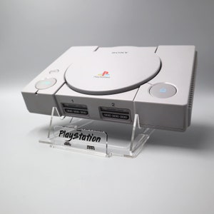 Könnte beinhalten: Eine weiße Sony Playstation Konsole mit einem klaren Acryl-Ständer. Die Konsole hat ein schwarzes PlayStation-Logo und zwei Speicherkartensteckplätze. Der Ständer hat ein schwarzes "Playstation"-Logo.