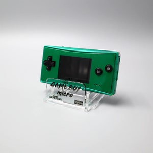 Könnte beinhalten: Eine grüne Game Boy Micro Handheld-Videospielkonsole auf einem durchsichtigen Acryl-Ständer. Die Konsole hat schwarze Knöpfe und einen schwarzen Bildschirm. Der Ständer trägt die Aufschrift "GAME BOY micro".
