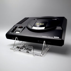 Può includere: Una console per videogiochi Sega Mega Drive a 16 bit nera su un supporto in acrilico trasparente. La console ha un design nero e argento con il testo "MEGA DRIVE SEGA" stampato sul davanti. Il supporto ha il testo "SEGA MEGA DRIVE" stampato su di esso.