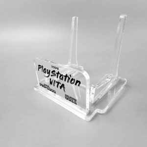 Può includere: Un supporto in acrilico trasparente per una console di gioco portatile Playstation Vita. Il supporto ha due supporti verticali e una base con il testo "TYPE Playstation VITA MODEL PCH-1000 RELEASE 2011" stampato su di esso.