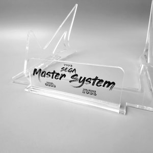 Puede incluir: Un expositor de acrílico transparente para una consola de videojuegos Sega Master System. El expositor presenta el texto "Type Sega Master System Model 3000 Release 1986".