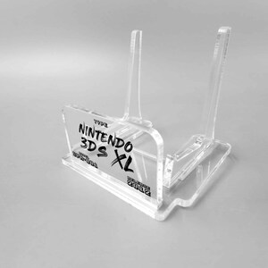 Könnte beinhalten: Klarer Acryl-Ständer für eine Nintendo 3DS XL-Konsole. Der Ständer trägt ein Etikett mit der Aufschrift "TYPE NINTENDO 3DS XL  MODEL  CTR-S-001 RELEASE 2012".