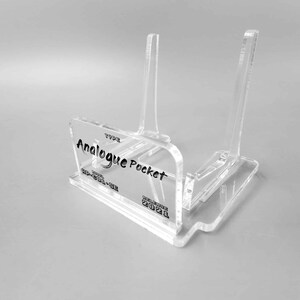 Könnte beinhalten: Klarer Acryl-Ständer für ein Handheld-Gaming-Gerät. Der Ständer trägt den Text "Analogue Pocket" und "Release 2021".