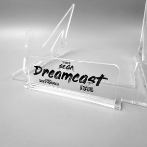 Puede incluir: Un soporte de exhibición de acrílico transparente para una consola Sega Dreamcast. El soporte tiene una etiqueta que dice "Type Sega Dreamcast Model HKT-3030 Release 1998".