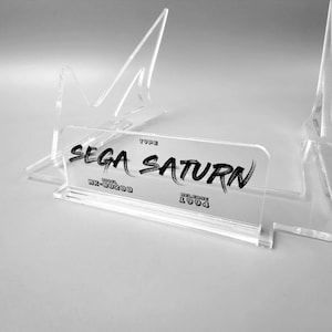 Könnte beinhalten: Ein transparenter Acryl-Präsentationsständer für eine Sega Saturn-Videospielkonsole. Der Ständer trägt ein Etikett mit der Aufschrift "TYPE SEGA SATURN MODEL MK-80200 RELEASE 1994".