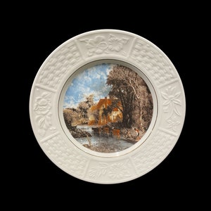 May include: A white ceramic plate with a raised rim and a woven pattern. The center of the plate features a printed image of a landscape with a river, trees, and a house.