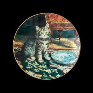 May include: A decorative ceramic plate featuring a kitten with green eyes sitting on a patterned rug. The plate has a blue and white bowl with a floral design on the rug.