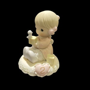 May include: A porcelain figurine of a young girl holding a block with the number "2" on it. She is sitting on a cloud-shaped base with a heart-shaped decoration that also has the number "2" on it. The figurine is painted in soft pink, white, and yellow tones.