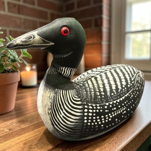 May include: A decorative loon figurine with a black head, neck, and back, and a white and black patterned body. The loon has red eyes and a detailed beak. The figurine is displayed on a wooden surface.