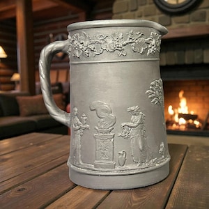 May include: A pewter-colored pitcher with a large handle and a spout. The pitcher is decorated with relief carvings of figures, trees, and grapevines. The pitcher is sitting on a wooden table.