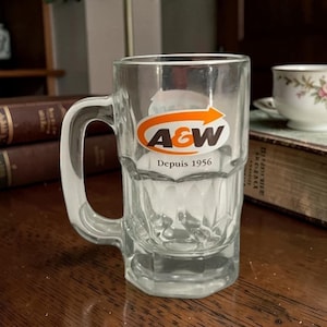 May include: Clear glass A&W mug with a handle. The mug features the A&W logo in orange and white with an arrow design, and the text "Depuis 1956". The mug is on a wooden surface.