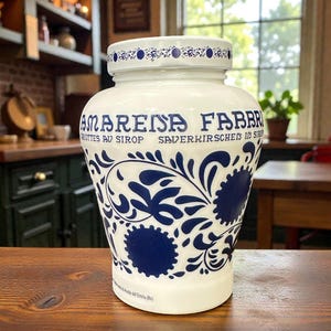 May include: A white ceramic jar with a blue floral design and text. The jar has a wide base and a narrower neck with a lid. The text includes the words "AMARENA FABBRI" and other text in multiple languages. The jar is sitting on a wooden surface.